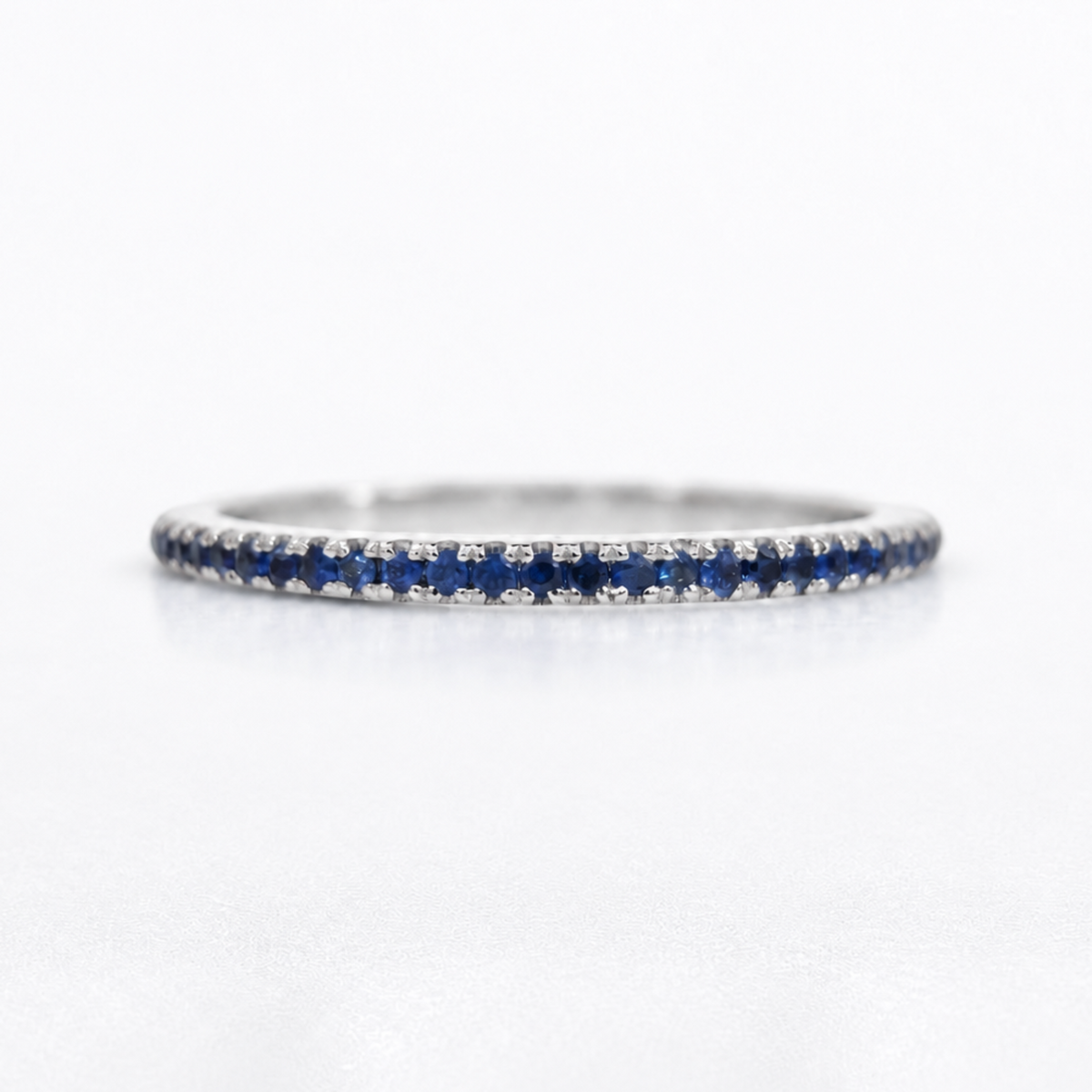 A thin silver eternity ring featuring a continuous row of deep blue round-cut sapphires on one side and small clear stones on the other, set against a clean white background.