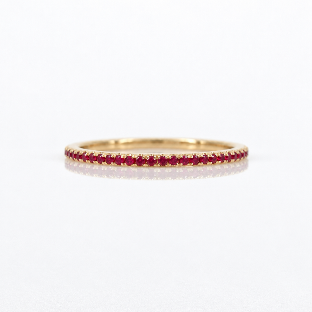 A thin yellow gold eternity ring set with a continuous row of small round-cut red rubies, photographed on a clean white reflective surface.