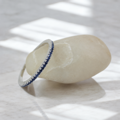 A thin silver band ring with a row of dark blue round blue sapphires, displayed on a smooth beige stone against a light marble surface with soft window-light shadows.