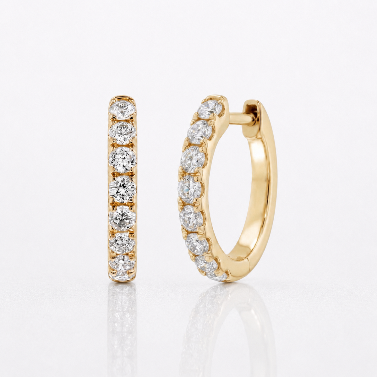 A pair of gold huggie hoop earrings, one shown front-facing and the other angled to show the side profile, both adorned with a row of round-cut clear diamonds, set against a clean white background with a soft reflection.