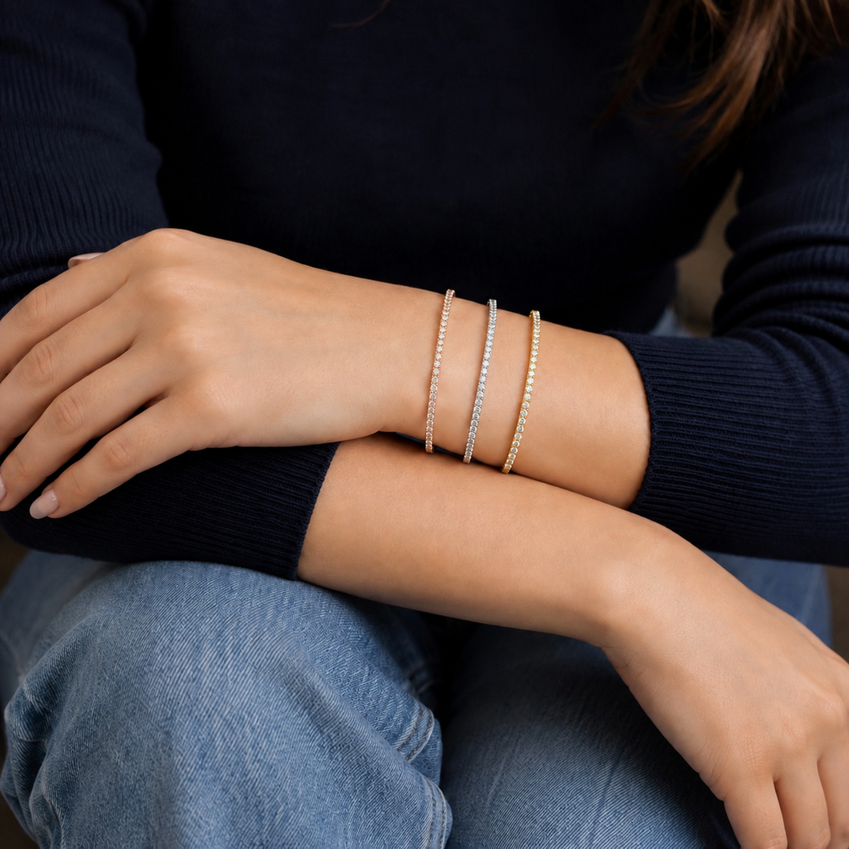 Three delicate bangle bracelets with pavé-set diamonds worn on a person's wrist, shown against a dark navy ribbed sweater and blue jeans.
