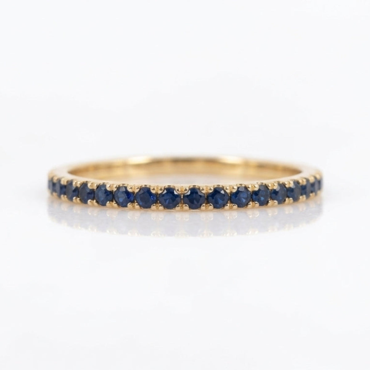 A thin yellow gold eternity ring set with a row of round-cut dark blue sapphires, shown on a clean white reflective surface.