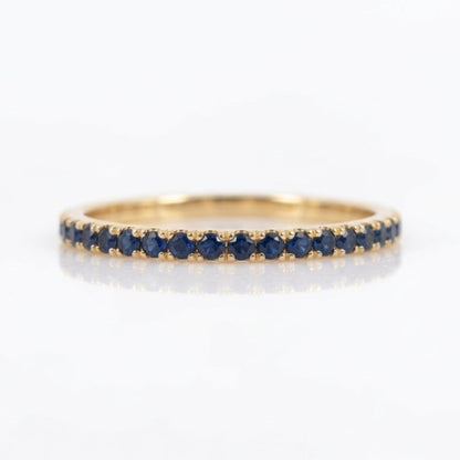 A thin yellow gold eternity ring set with a row of round-cut dark blue sapphires, shown on a clean white reflective surface.