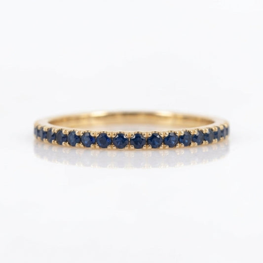 A thin yellow gold eternity ring set with a row of round-cut dark blue sapphires, shown on a clean white reflective surface.
