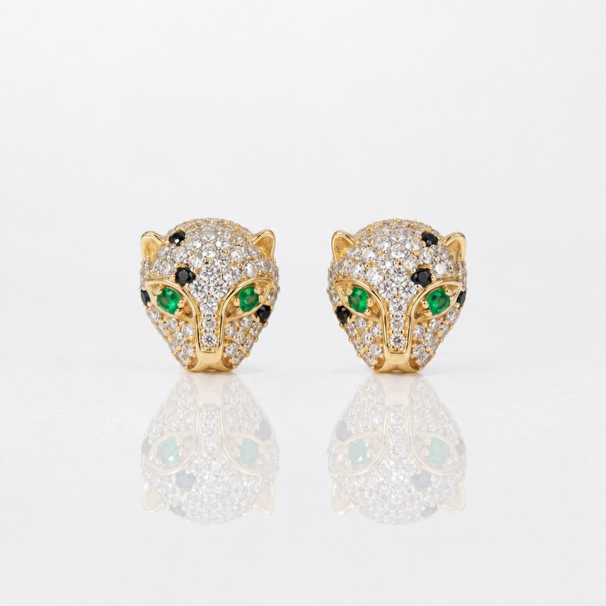 A pair of gold leopard head stud earrings paved with clear diamonds, featuring green emerald-cut gemstones for the eyes and small black stones for the eyebrows, displayed on a reflective white surface.