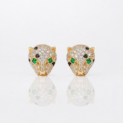 A pair of gold leopard head stud earrings paved with clear diamonds, featuring green emerald-cut gemstones for the eyes and small black stones for the eyebrows, displayed on a reflective white surface.
