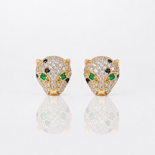 A pair of gold leopard head stud earrings paved with clear diamonds, featuring green emerald-cut gemstones for the eyes and small black stones for the eyebrows, displayed on a reflective white surface.