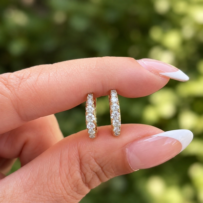 A close-up of a hand with a French manicure holding a pair of gold half-ear hoop earrings set with small clear diamonds  against a blurred green outdoor background.
