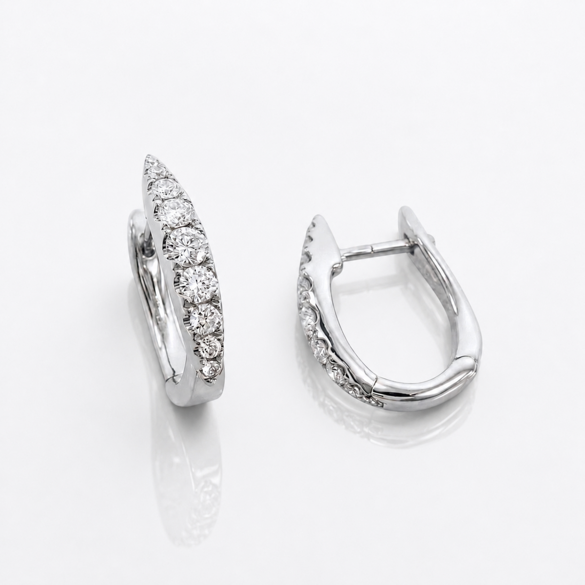 A pair of silver huggie hoop earrings with pavé-set round diamonds, shown from two angles on a reflective white surface.