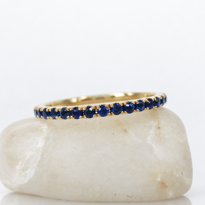 A yellow gold eternity ring set with round-cut deep blue sapphires, displayed on a smooth white stone against a clean white background.