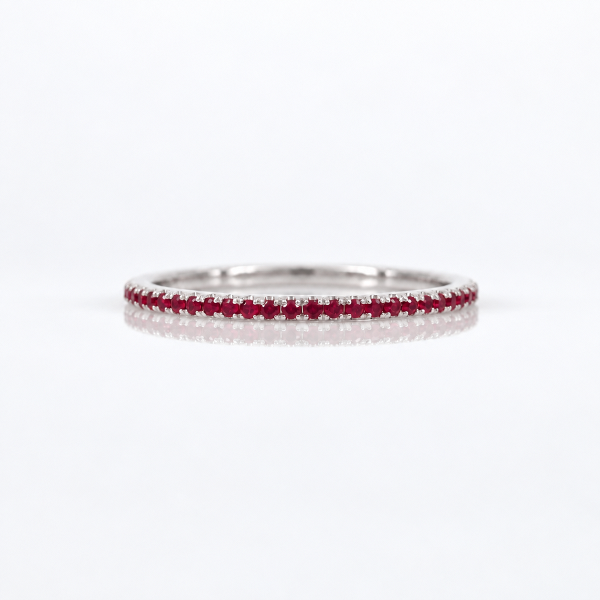A thin silver eternity ring set with a single row of round-cut red rubies, photographed on a clean white reflective surface.