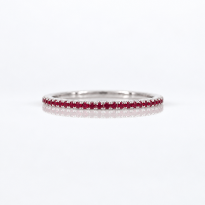 A thin silver eternity ring set with a single row of round-cut red rubies, photographed on a clean white reflective surface.