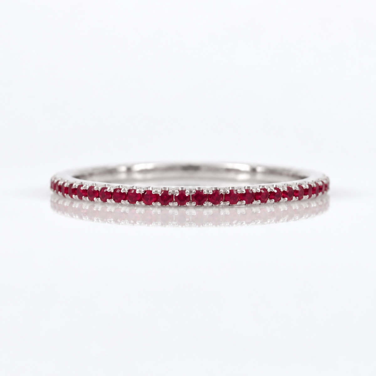 A thin silver-tone eternity ring set with a continuous row of small round-cut red rubies, photographed on a clean white background with a soft reflection.