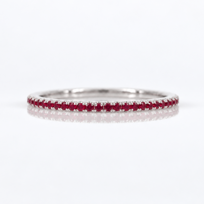 A thin silver-tone eternity ring set with a continuous row of small round-cut red rubies, photographed on a clean white background with a soft reflection.