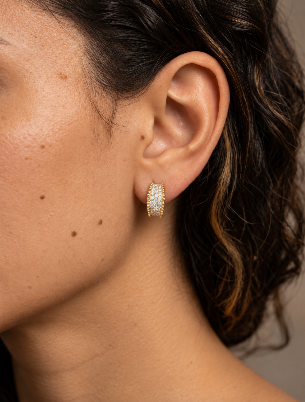 Close-up of a model wearing a 12 mm yellow gold natural diamond pavé huggie hoop earring with multiple rows of round diamonds and polished gold beaded edges.