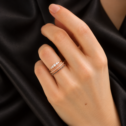 A close-up of a hand wearing three delicate rings against a black satin fabric background. The rings are thin rose gold bands, with the center ring featuring small clear stones.