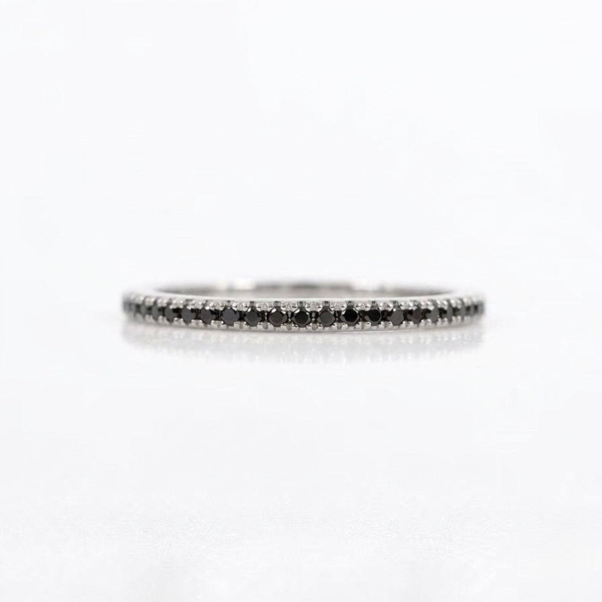 A thin silver-tone eternity ring set with small black diamonds, photographed on a clean white background with a soft reflection.