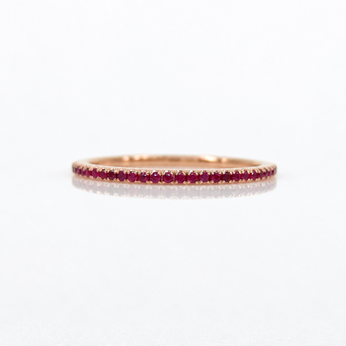 A thin rose gold eternity ring paved with small red rubies, shown on a clean white reflective surface.