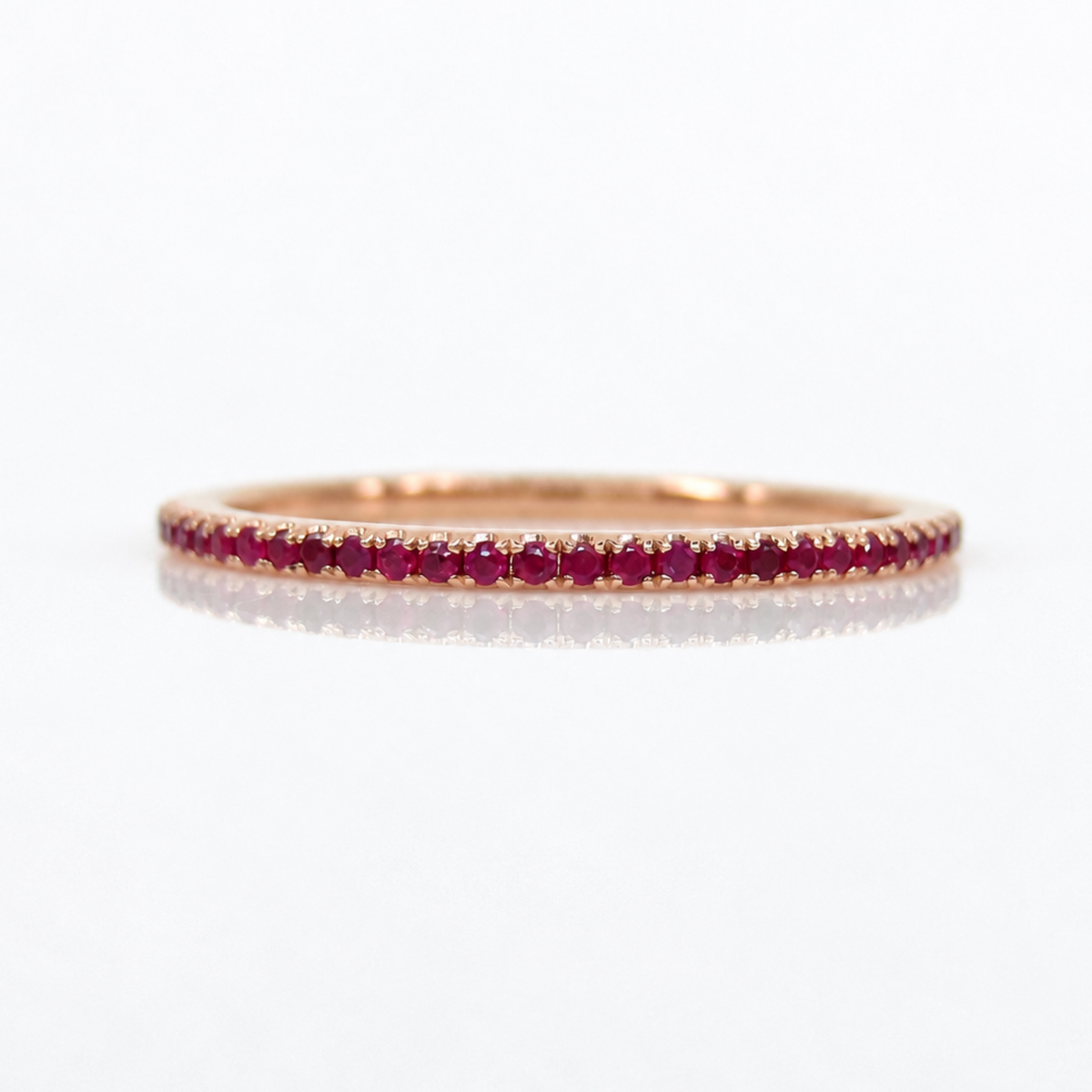 A rose gold eternity ring set with a row of round-cut red rubies, shown on a clean white reflective surface.