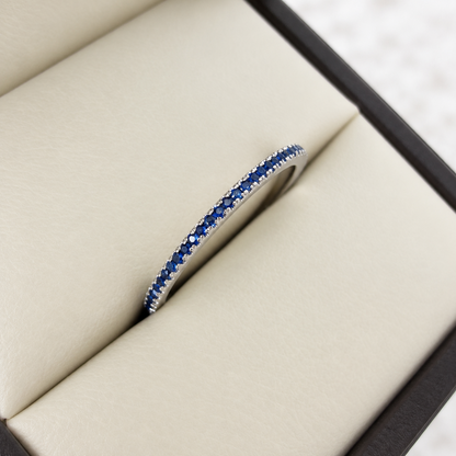 A silver eternity ring with a double row of round-cut blue sapphires, displayed inside a cream-colored jewelry box.