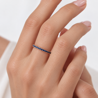 A close-up of a hand wearing a delicate rose gold eternity ring set with small round blue sapphires, shown against a clean white background.