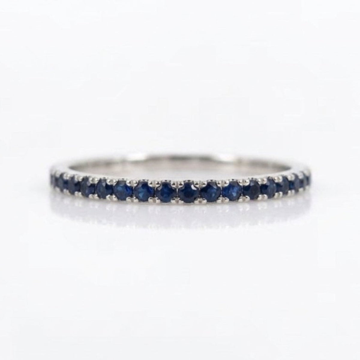 A thin silver eternity ring set with round-cut dark blue sapphires, shown on a clean white reflective surface.