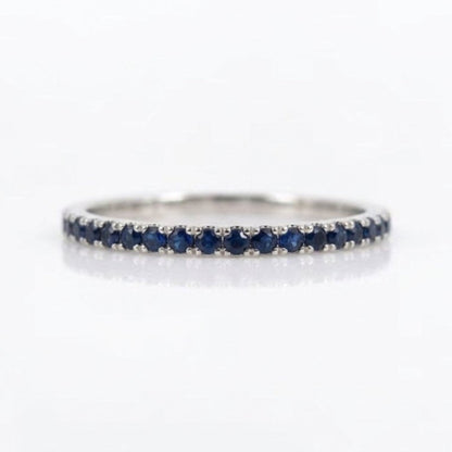 A thin silver eternity ring set with round-cut dark blue sapphires, shown on a clean white reflective surface.