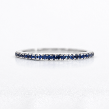 A thin silver eternity ring featuring a continuous row of deep blue round-cut sapphires on one side and small clear stones on the other, set against a clean white background.