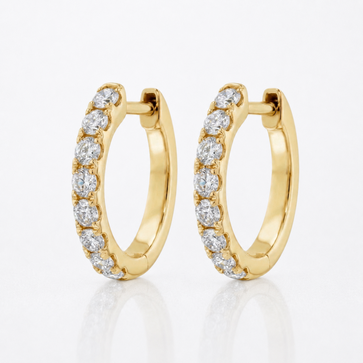 A pair of gold huggie hoop earrings set with round clear diamonds, displayed on a clean white reflective surface.