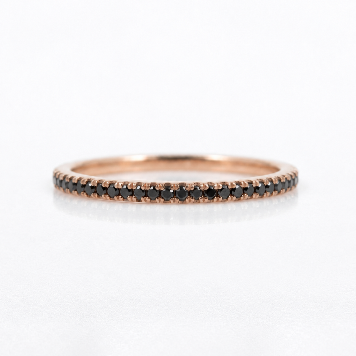 A thin rose gold eternity ring set with small round black diamonds, shown on a clean white background with a soft reflection.
