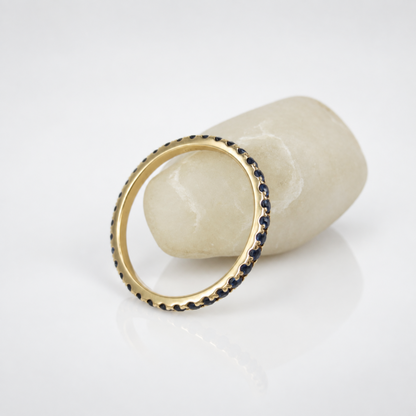 A gold ring with a border of small dark blue stones, displayed on a white surface next to a light-colored stone prop.