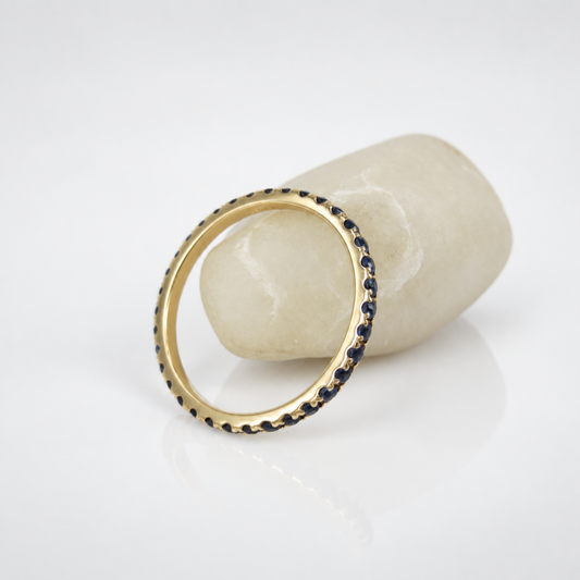 A gold ring with a border of small dark blue stones, displayed on a white surface next to a light-colored stone prop.