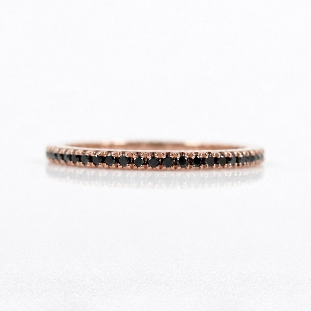 A delicate rose gold eternity ring featuring a row of small black diamonds, photographed on a clean white reflective surface.