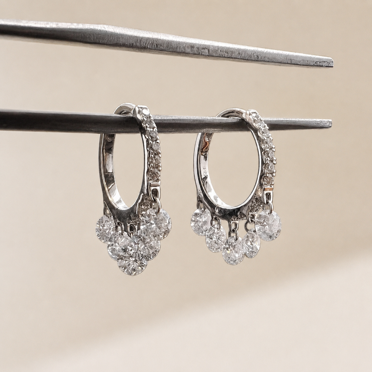 A pair of silver hoop earrings with dangling clear diamonds clusters, held by metal tweezers against a soft beige background.