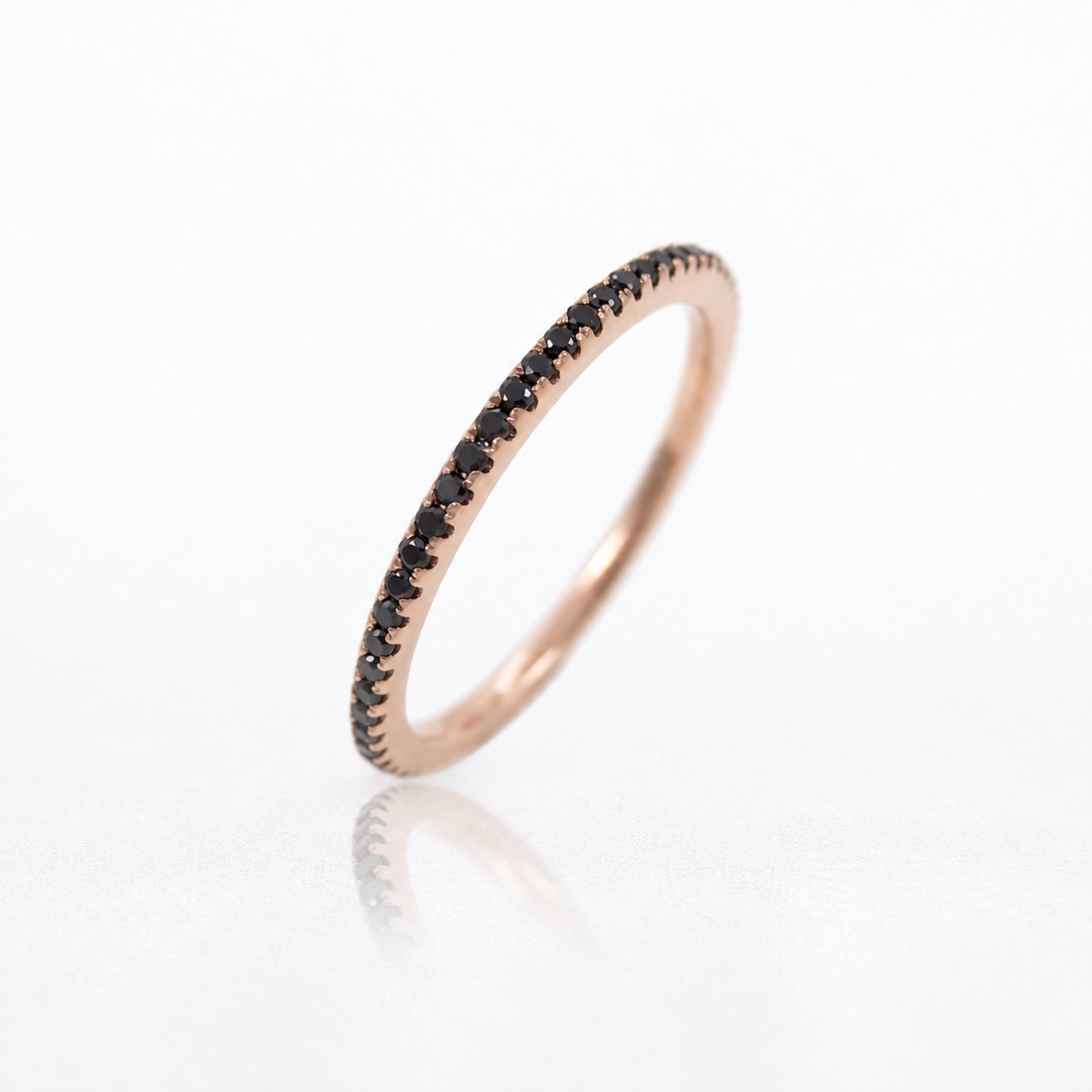 A rose gold eternity ring featuring a single row of small round-cut black diamonds, photographed on a clean white reflective surface.