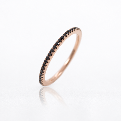 A rose gold eternity ring featuring a single row of small round-cut black diamonds, photographed on a clean white reflective surface.