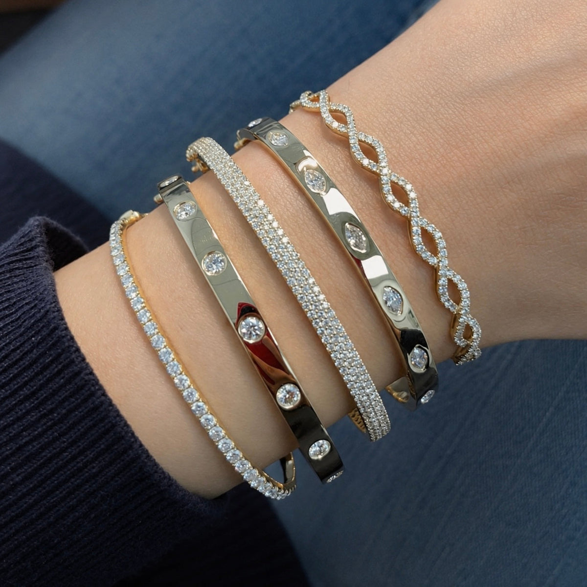 A close-up of a person's wrist wearing five stacked bangle bracelets. The bracelets include gold, silver, and rose gold metals, some paved with clear stones and others featuring round inset stones.