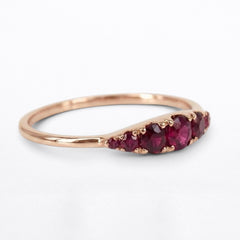 A rose gold ring featuring a curved row of oval deep red rubies, photographed on a clean white background.