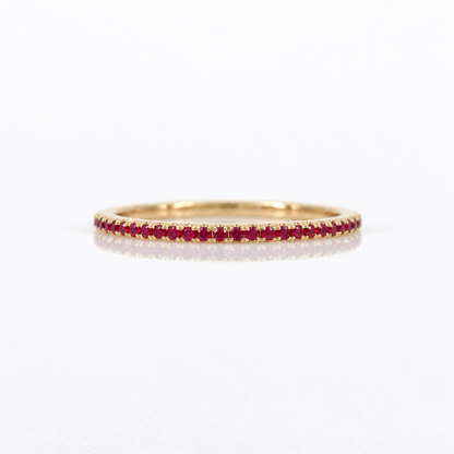 A thin yellow gold eternity ring set with a continuous row of small round-cut red rubies, photographed on a clean white reflective surface.
