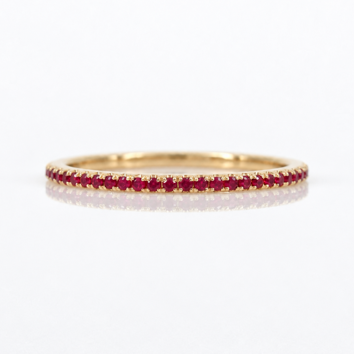 A thin yellow gold eternity ring set with a row of round-cut red rubies, shown on a clean white background with a soft reflection.