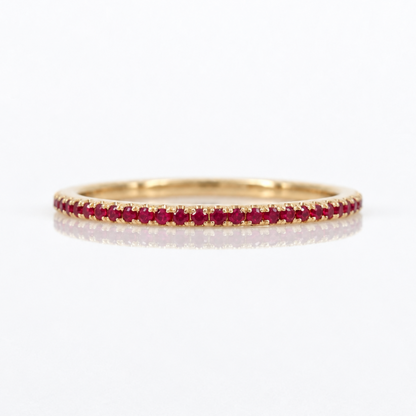 A thin yellow gold eternity ring set with a row of round-cut red rubies, shown on a clean white background with a soft reflection.