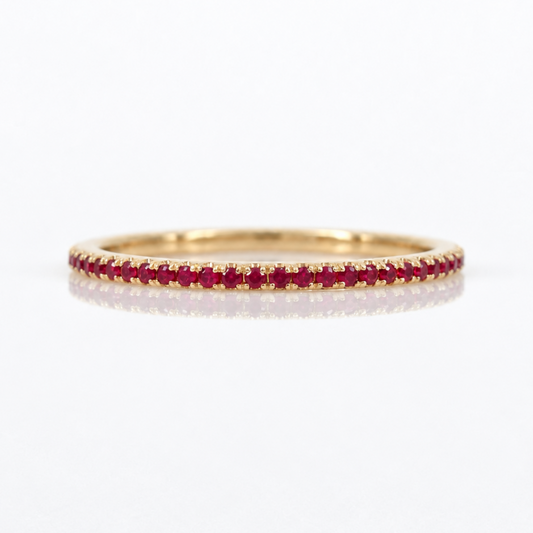 A thin yellow gold eternity ring set with a row of round-cut red rubies, shown on a clean white background with a soft reflection.