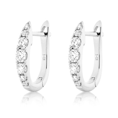 A pair of silver huggie hoop earrings with a row of clear round-cut diamonds  on the front, shown on a white background with a soft reflection below.