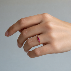 A close-up of a hand wearing a delicate gold ring set with graduated seven stone red rubies, shown against a soft light gray background.