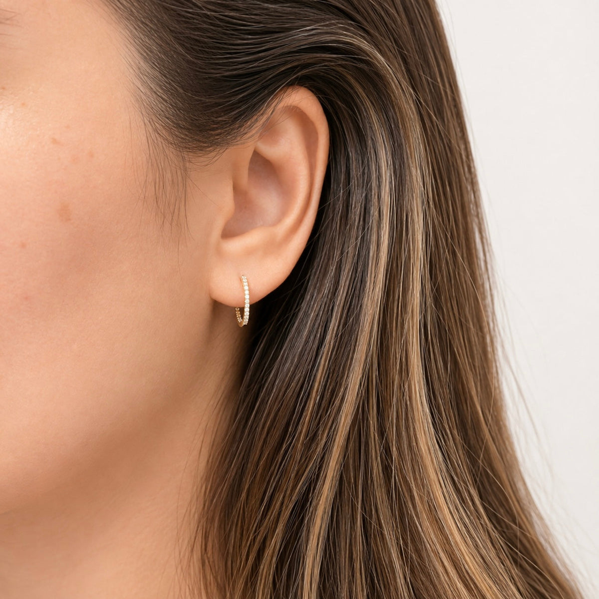 A close-up of a woman's ear wearing a delicate gold huggie hoop earring with tiny pavé-set diamonds, against a clean white background.