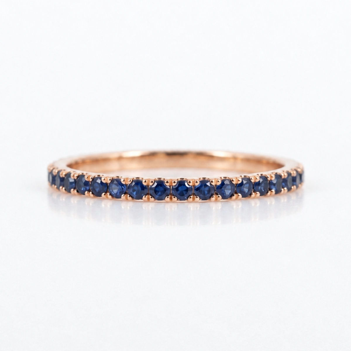 A rose gold eternity ring set with a row of round-cut deep blue sapphires, shown on a clean white background with a soft reflection.