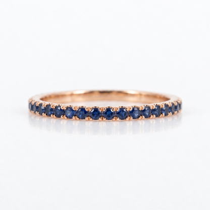 A rose gold eternity ring set with a row of round-cut deep blue sapphires, shown on a clean white background with a soft reflection.