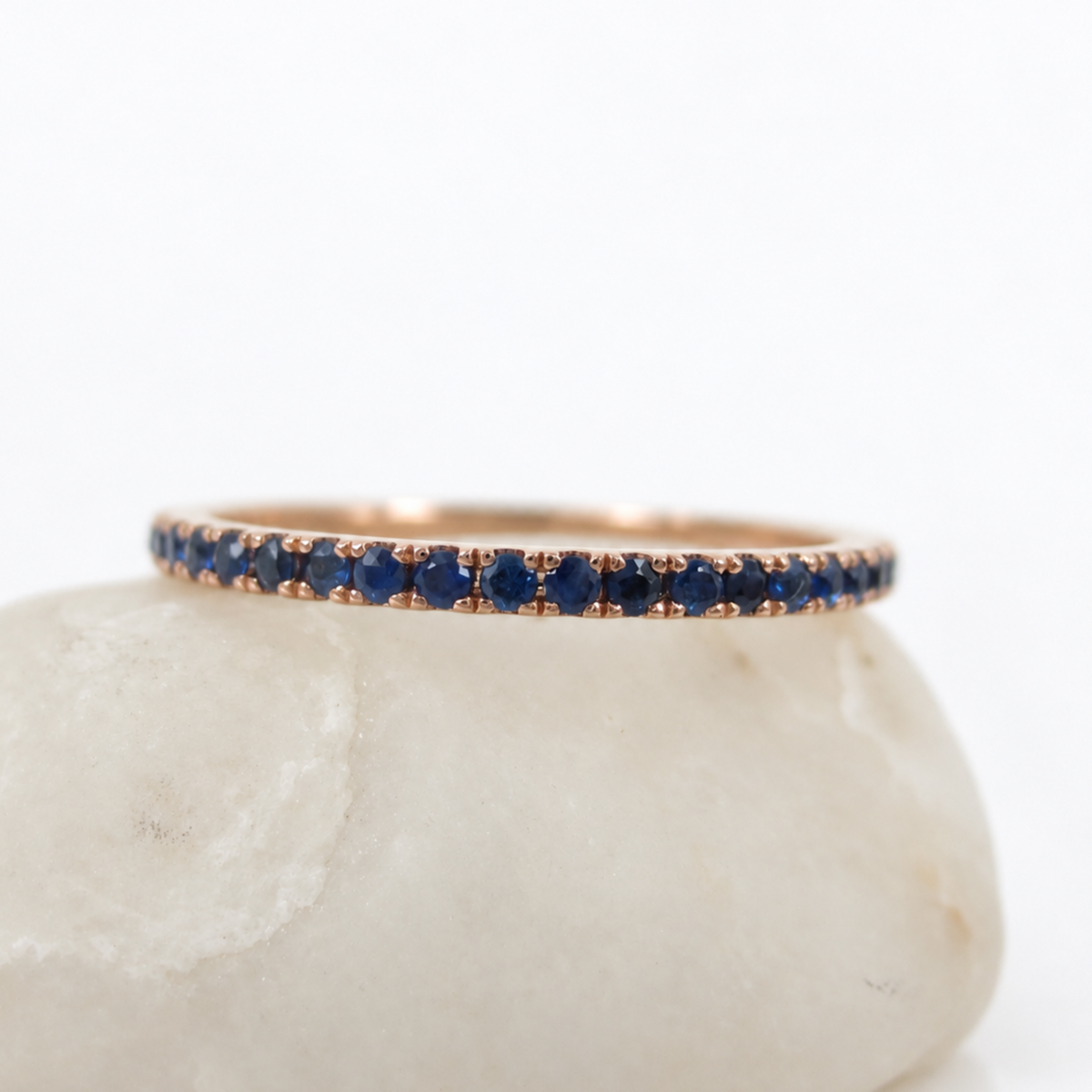A thin rose gold eternity ring set with round blue sapphires, displayed on a smooth white stone against a white background.