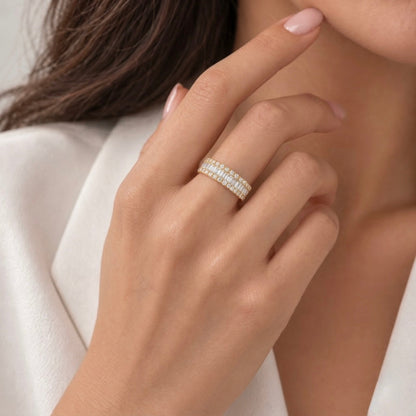 A close-up of a model's hand wearing a delicate double-row diamond eternity ring in yellow gold, set against a soft white blazer and neutral background.