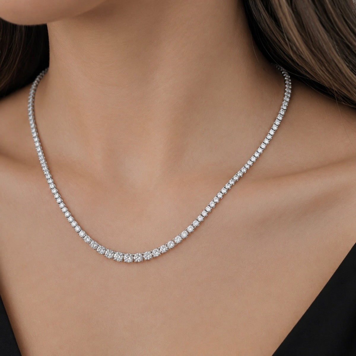 A close-up of a woman's neck and upper chest wearing a delicate silver tennis necklace with small clear diamonds.